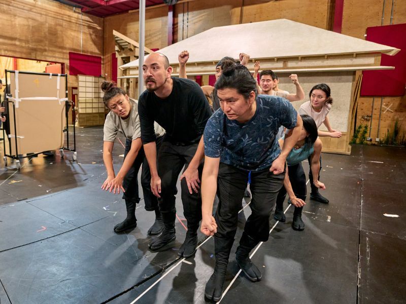 Image: cast members rehearsing, they appear to be bending down and focusing on something.