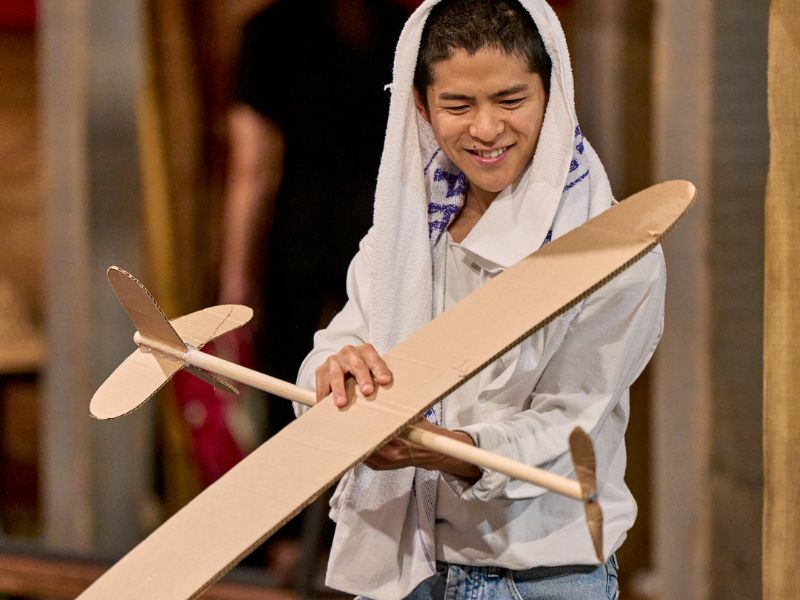 Image: a cast member holding a wooden airplane.