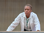 A person in a lab coat leans forward with a serious expression. They appear focused and concerned.