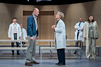 A dramatic scene features two individuals facing each other, while others observe. The setting is a minimalist stage with benches and a wooden backdrop.