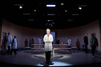 A person in a lab coat stands in the foreground, surrounded by others in a theatrical set. The scene is illuminated by dramatic lighting against a circular backdrop.