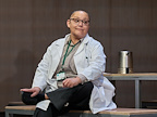 Naomi Wirthner (Hardiman) - The Doctor - wearing hospital scrubs and looking off into the distance.