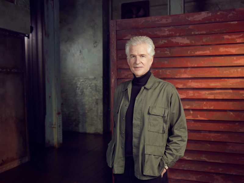 Image: Matthew Modine stood in front of an amber panelled background, he is looking at the camera and is wearing a green jacket.