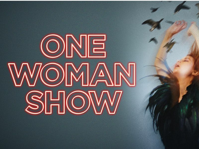 Text: One Woman Show, in neon red writing. Image: Liz Kingsman on a grey/navy background with her hands up, wearing black surrounded by birds.
