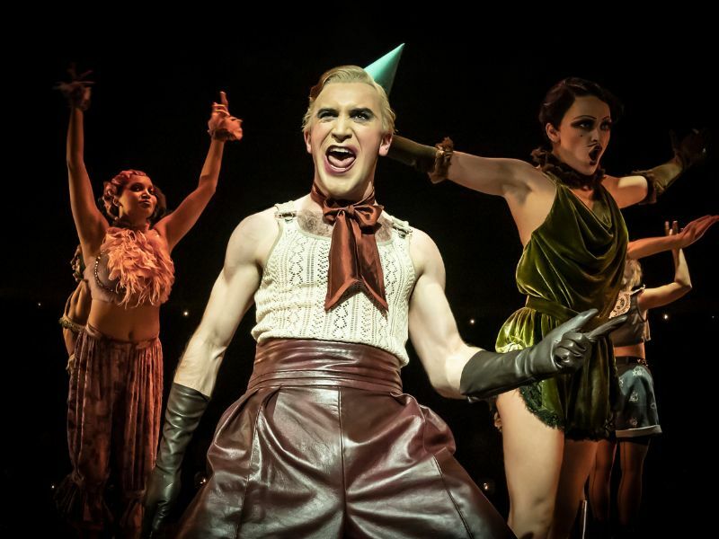 Callum Scott Howells as Emcee Cabaret. He is dressed in a party hat with pale makeup and a is wearing a white tank top, he is smiling chaotically.