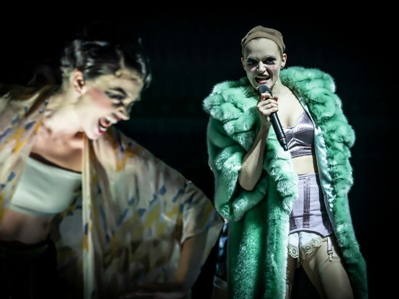 Madeline Brewer as Sally Bowels, she is wearing a fluffy green coat and pale makeup and is singing into a microphone.