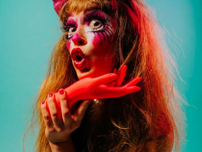 Image: headshot of Tara Boom, holding an orange hand and wearing clown makeup. 