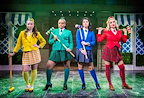 Four individuals stand confidently in colorful outfits on a stage, each holding a croquet mallet. The vibrant setting features greenery and decorative elements in the background.
