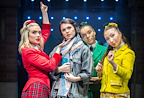 Four individuals pose dramatically on stage, dressed in colorful costumes. They exhibit confident expressions against a backdrop of stage lights.