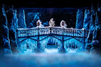 A dramatic stage scene depicts a snowy bridge surrounded by icy formations and mist. Two figures stand on the bridge, immersed in a magical winter setting.