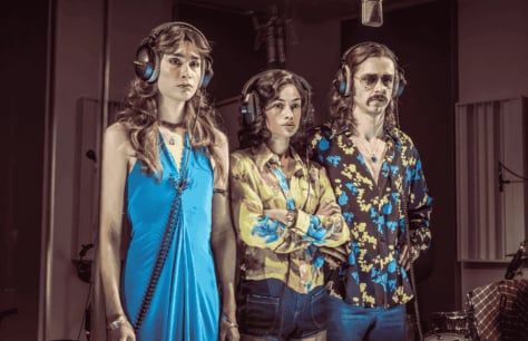 Three musicians in 1970s outfits gather around a microphone in a recording studio. 